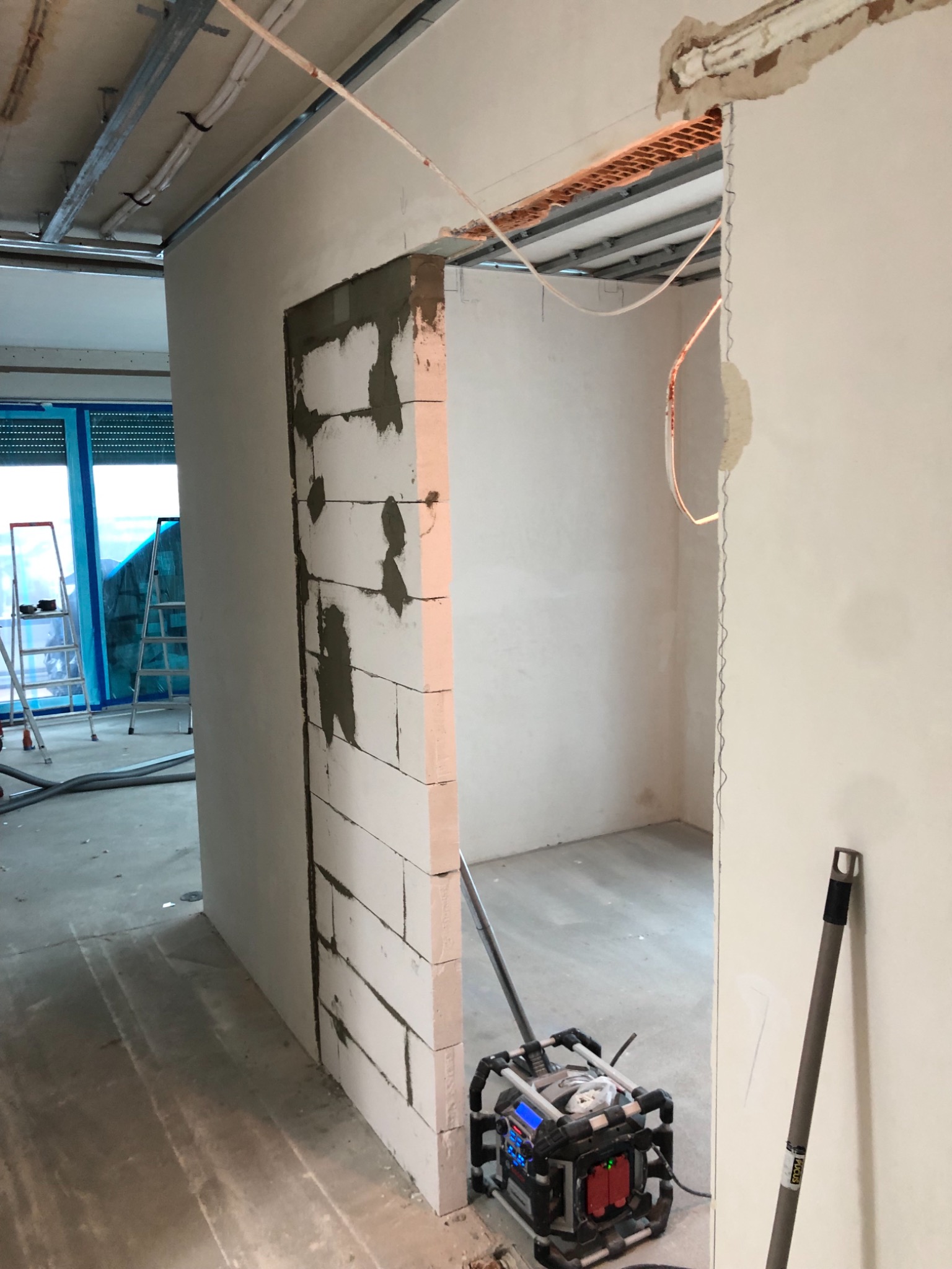 Wnętrze w trakcie budowy ściany z białych bloczków, widoczne metalowe profile pod sufitem, przewody elektryczne i przenośna lampa budowlana na podłodze.