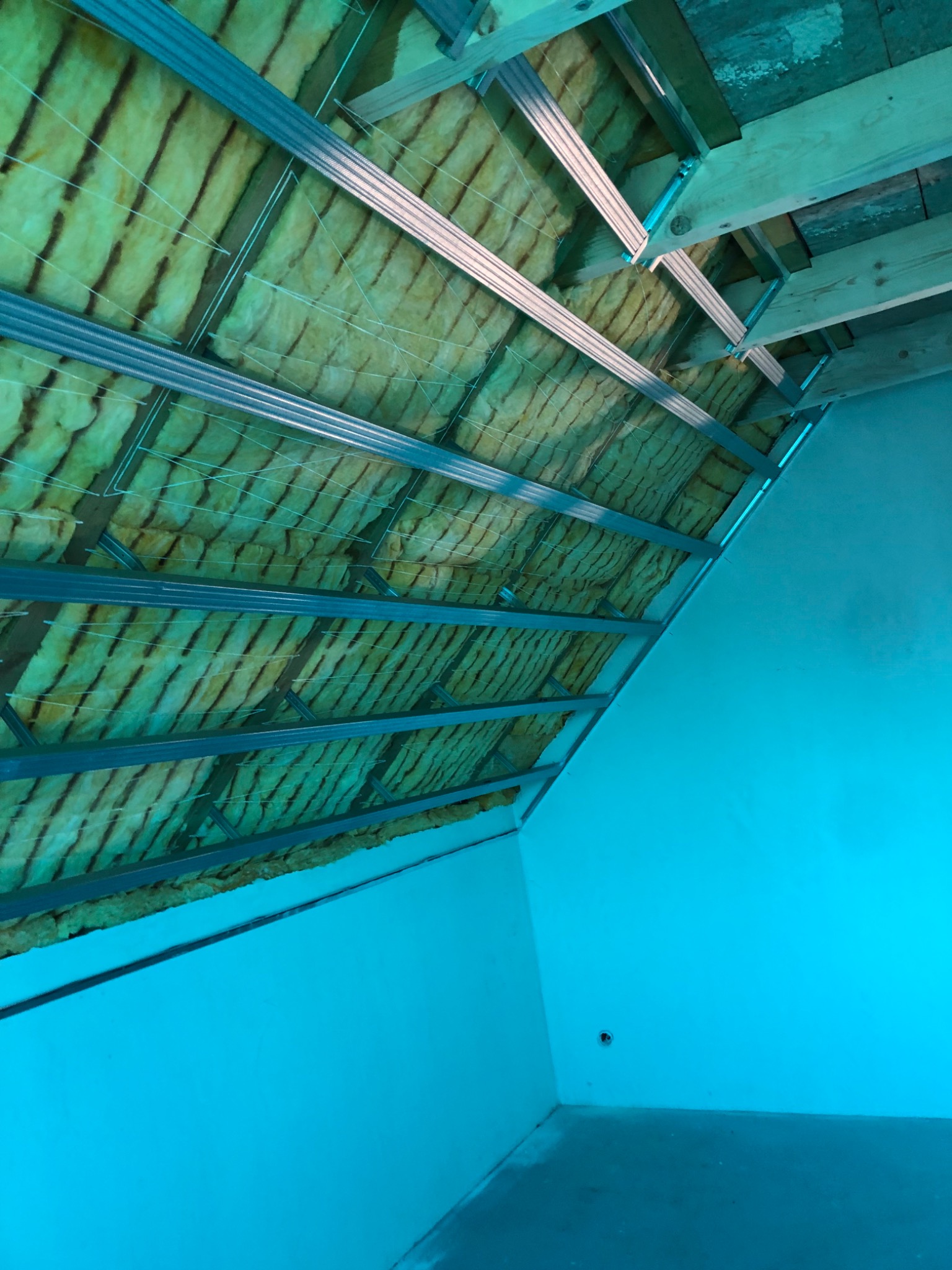 Wnętrze poddasza w trakcie izolacji: widoczne metalowe profile sufitowe, wełna mineralna zabezpieczona sznurkiem oraz fragment pomalowanej ściany w kolorze turkusowym.