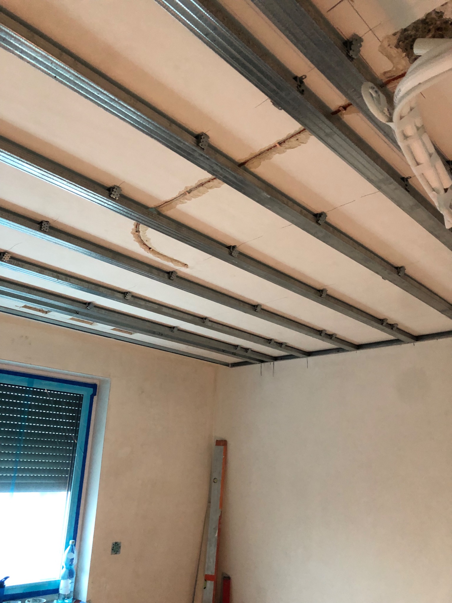 Montaż metalowej konstrukcji podwieszanego sufitu w pomieszczeniu z oknem i butelką wody, widoczne elementy instalacji elektrycznej.