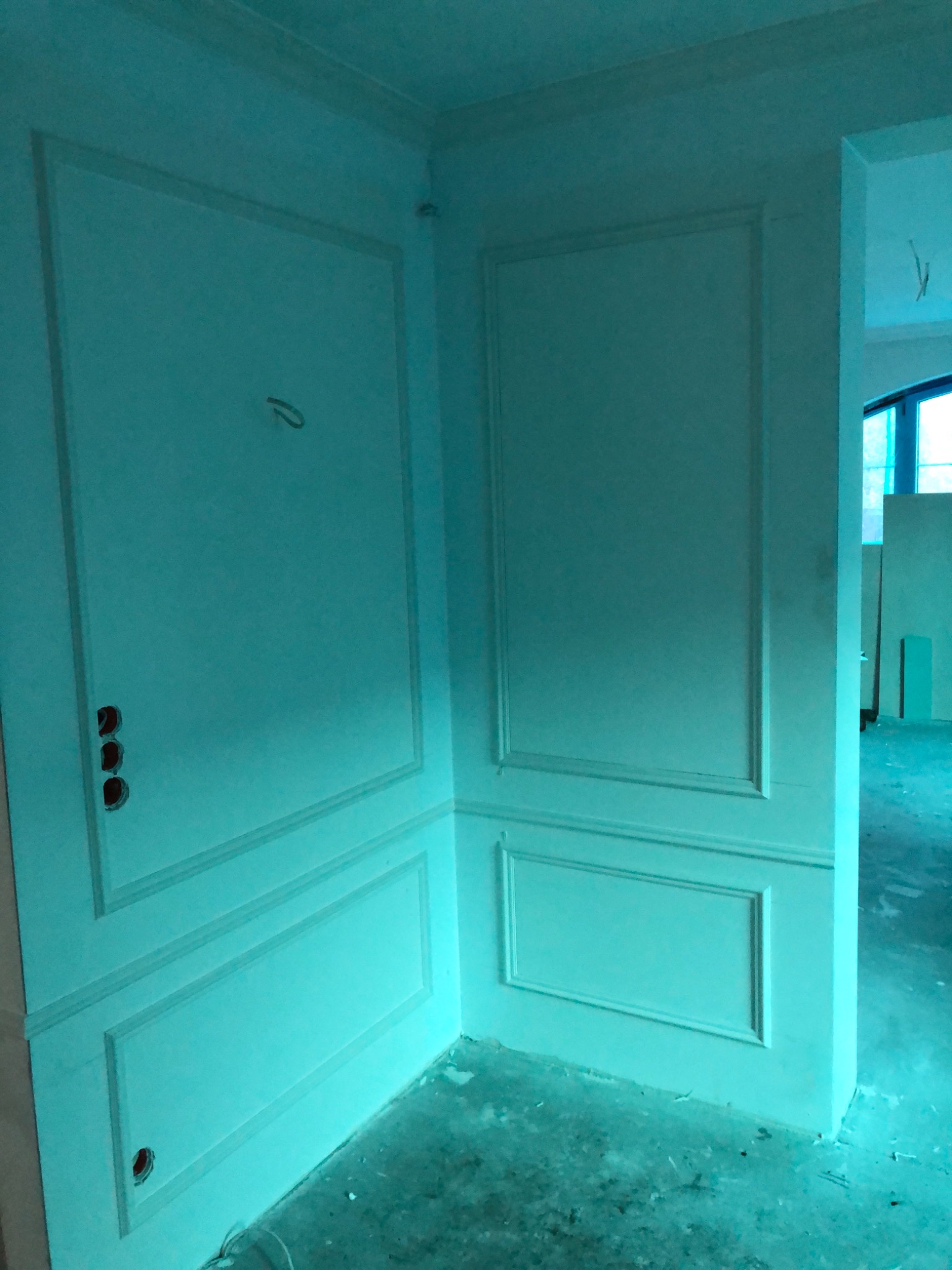 Fragment ściany w rogu pomieszczenia z dekoracyjnymi panelami ściennymi w trakcie prac wykończeniowych, widoczne otwory na gniazdka elektryczne i niedoskonałości podłoża.