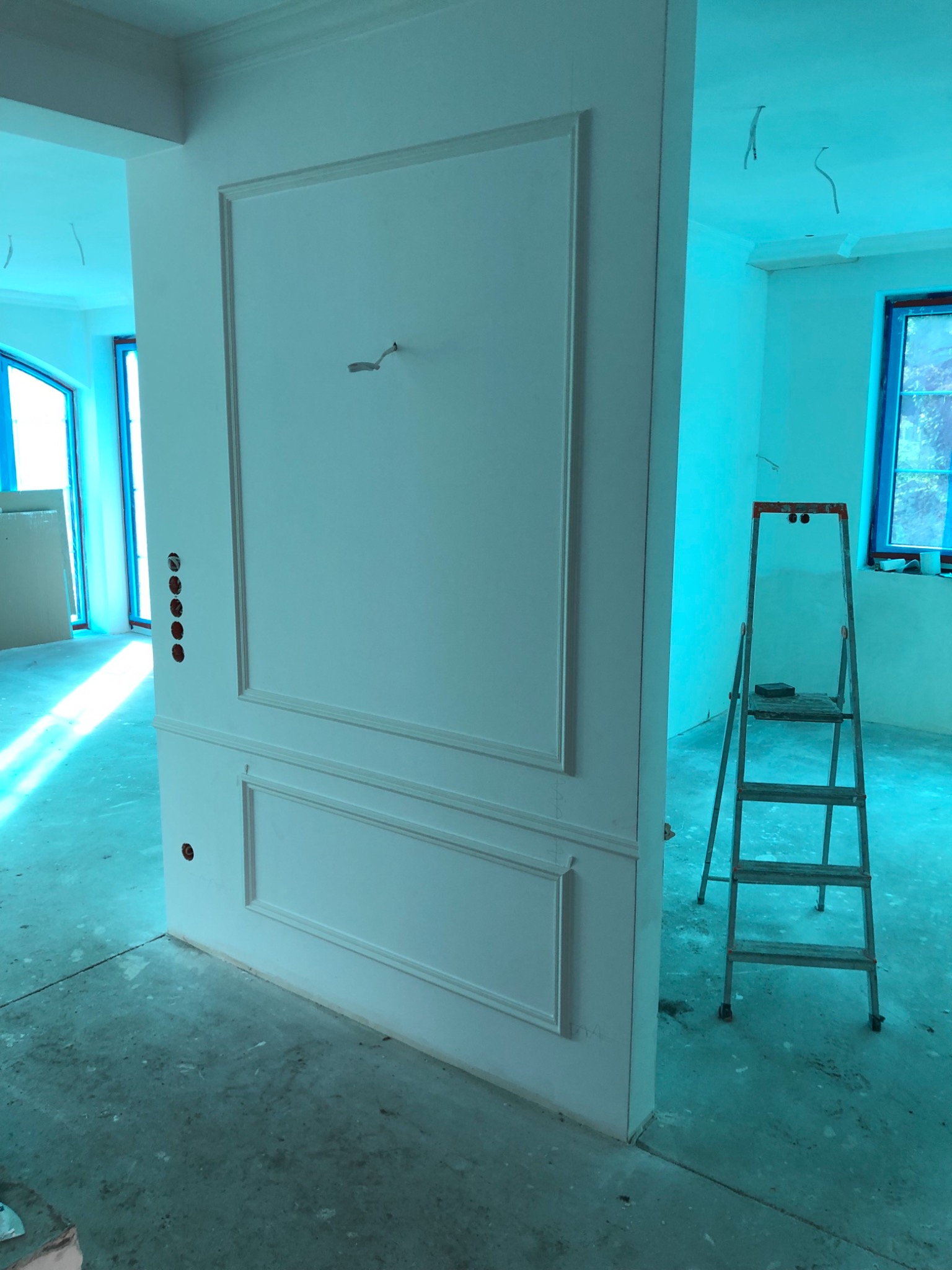 Ściana działowa z dekoracyjną sztukaterią i otworami na gniazdka elektryczne, widoczna drabina aluminiowa, surowy beton na podłodze i okno w tle, wnętrze w trakcie prac wykończeniowych.