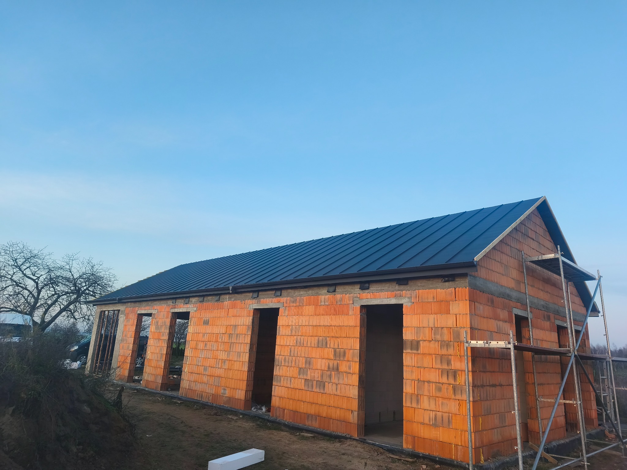 Budowa domu z czerwonej cegły w Głodnie, z zamontowanym ciemnym dachem. Widoczne rusztowania i otwory okienne/drzwiowe. Błękitne niebo w tle.