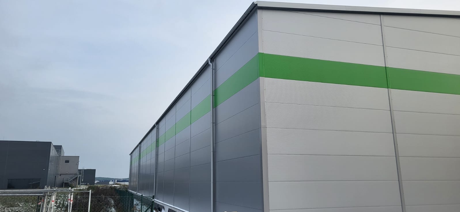 Budynek przemysłowy z płyt warstwowych w odcieniach szarości i bieli, z poziomym zielonym pasem. Widok z dołu, fragment nieba w tle. Nowoczesna architektura.