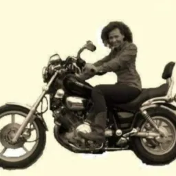 Kobieta z kręconymi włosami siedzi na czarnym motocyklu cruiser z oparciem, ubrana w ciemne spodnie i koszulę, uśmiechając się w stronę obiektywu.