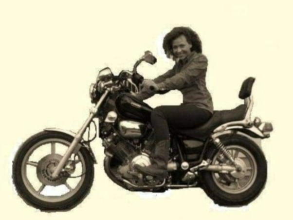 Kobieta z kręconymi włosami siedzi na czarnym motocyklu cruiser z oparciem, ubrana w ciemne spodnie i koszulę, uśmiechając się w stronę obiektywu.