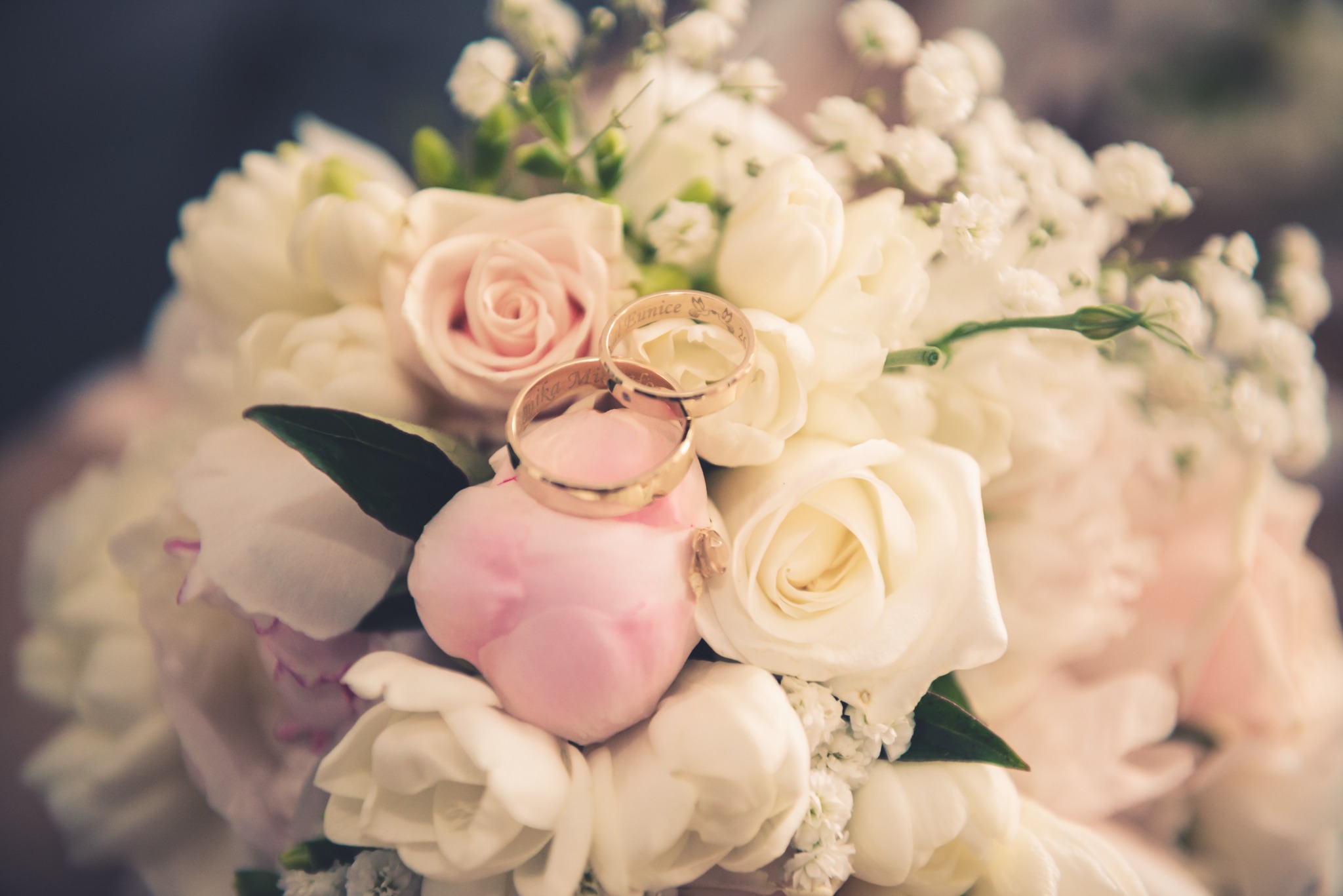 Dwa złote obrączki z wygrawerowanymi imionami leżą na bukiecie ślubnym z białych i różowych róż w Mińsku Mazowieckim.