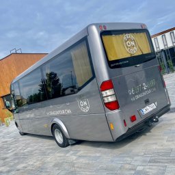 DRAGonTOUR - Szary minibus Mercedes-Benz Sprinter z logo 'Dragon Tour' na tylnej szybie i boku, zaparkowany na szarej kostce brukowej przed nowoczesnym budynkiem z drewnianą elewacją.