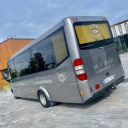 Szary minibus Mercedes-Benz Sprinter z logo 'Dragon Tour' na tylnej szybie i boku, zaparkowany na szarej kostce brukowej przed nowoczesnym budynkiem z drewnianą elewacją.