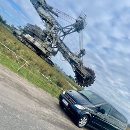 DRAGonTOUR - Czarny van Mercedes-Benz Viano na tle ogromnej koparki kołowej, widok z dołu, pochmurne niebo w tle.