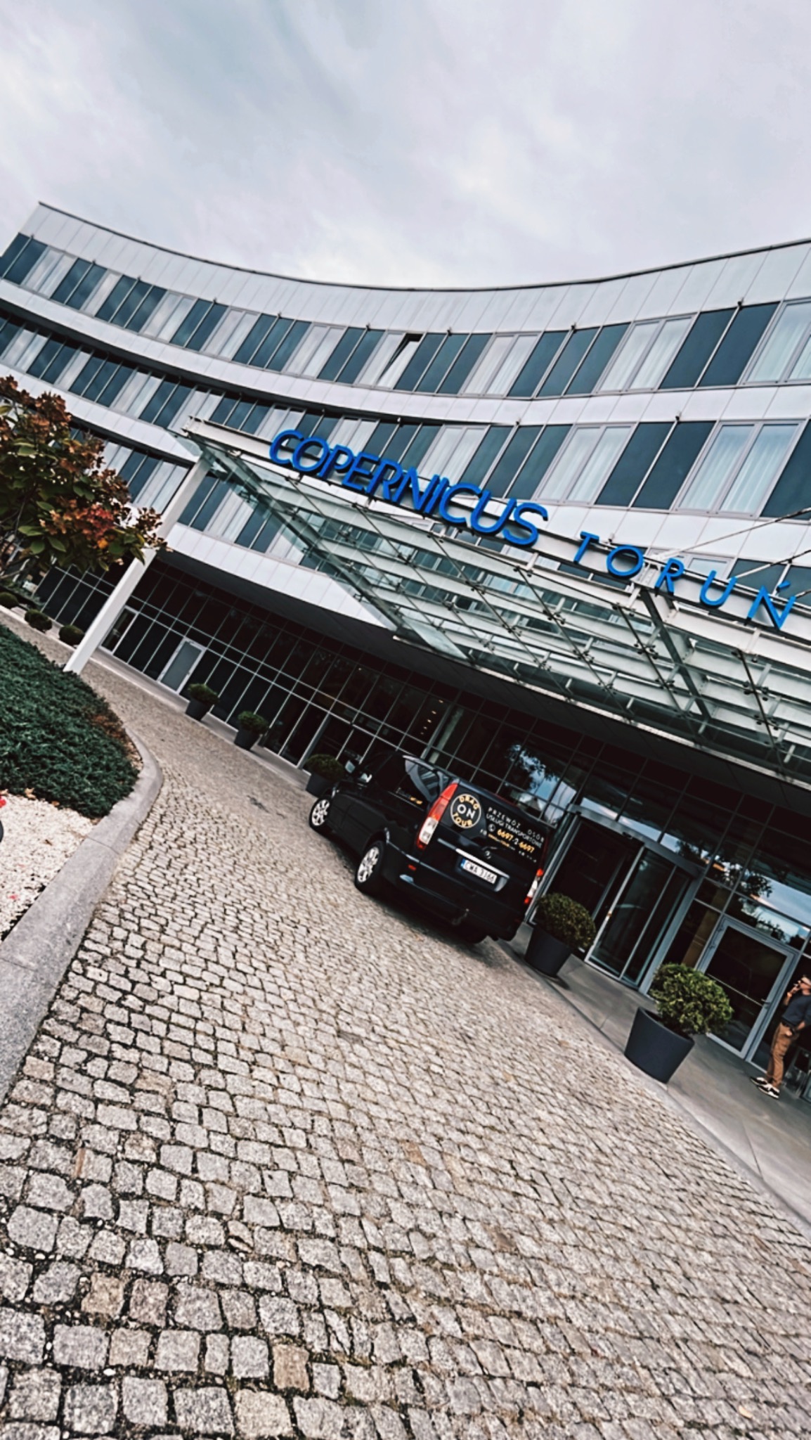 Czarny bus z reklamą przewozu osób zaparkowany przed budynkiem Copernicus Toruń, widok z perspektywy chodnika z kostki brukowej.