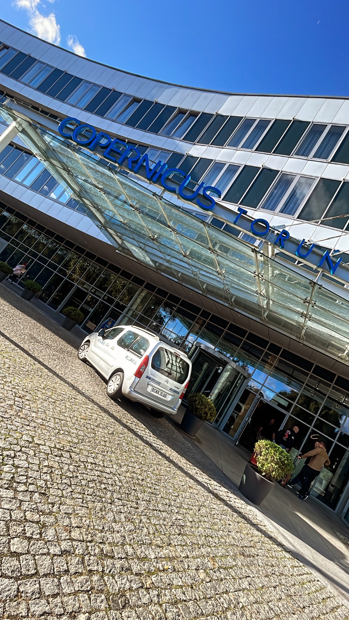 Srebrny samochód typu van zaparkowany przed nowoczesnym budynkiem biurowym Copernicus Forum z przeszklonym wejściem i niebieskim napisem, widok z perspektywy chodnika z kostki brukowej.