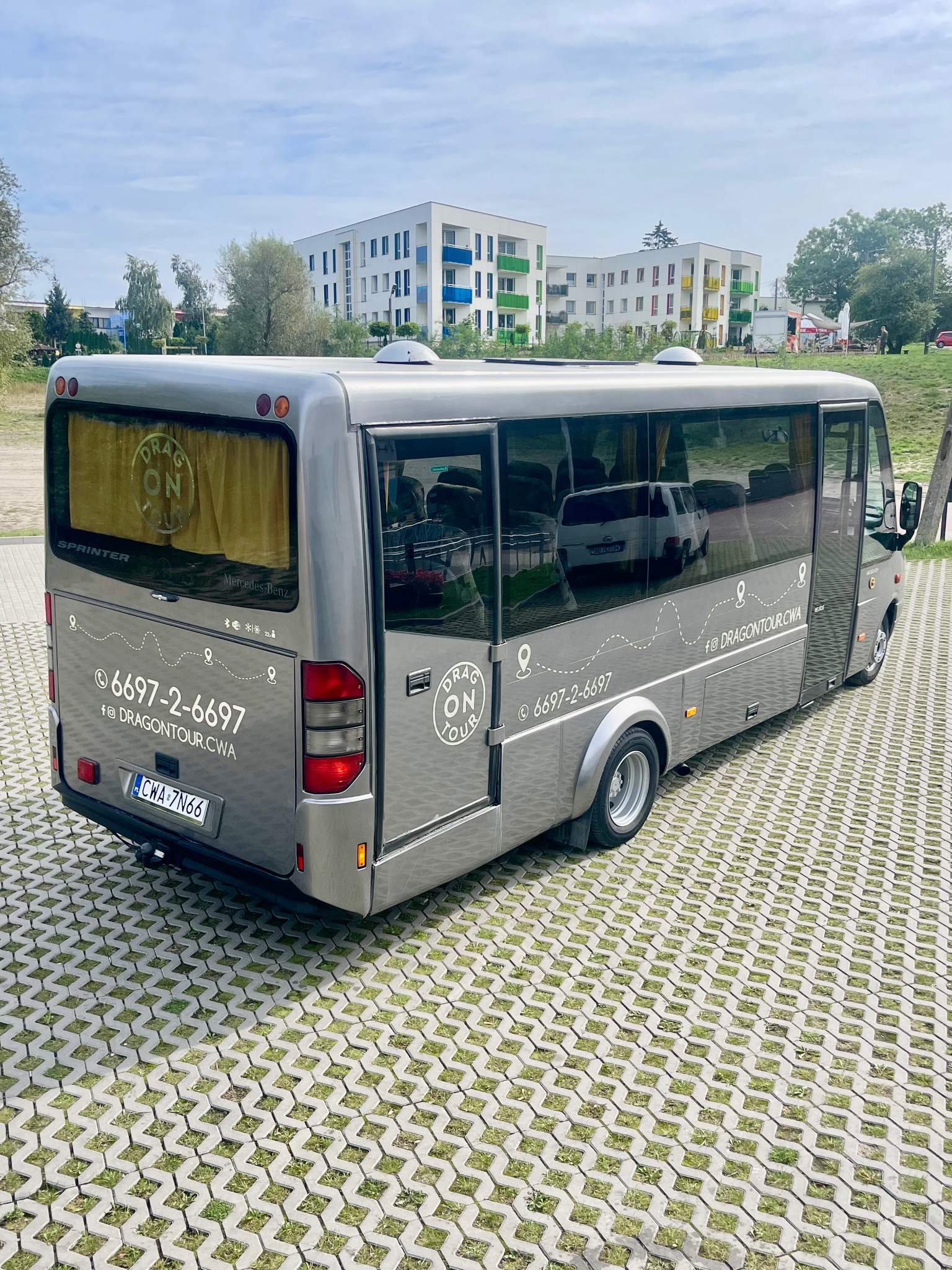 Szary bus Mercedes-Benz Sprinter z logo 'Dragon Tour' na parkingu z ażurowych płyt, w tle budynki mieszkalne z kolorowymi balkonami.
