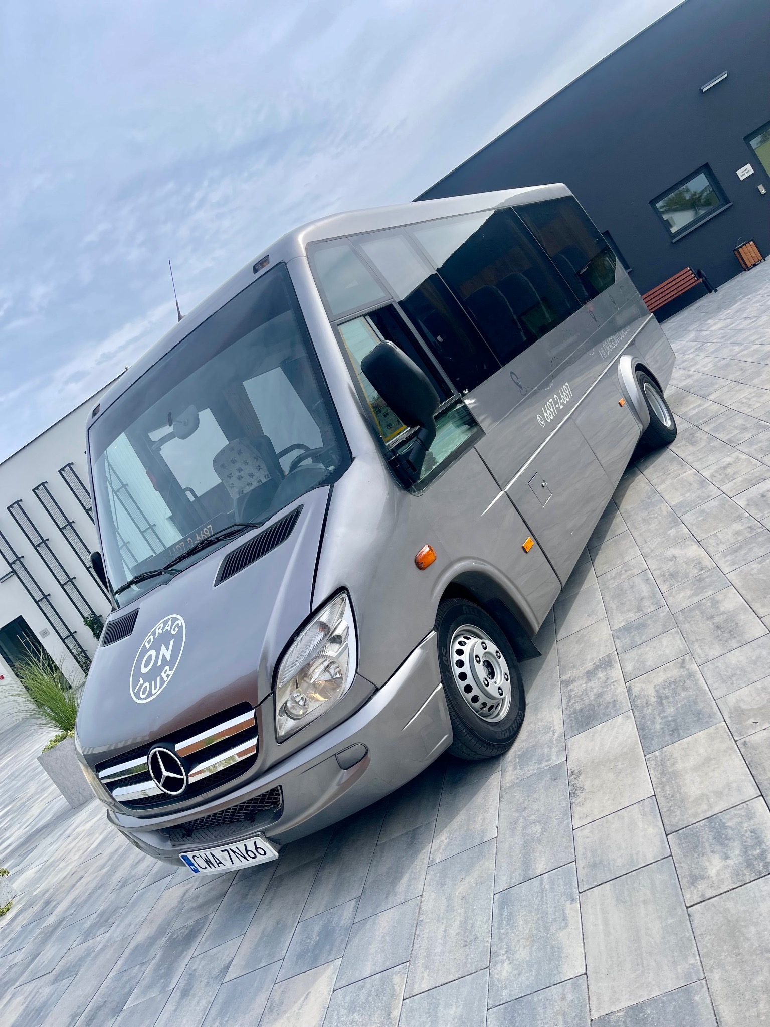 Szary bus marki Mercedes z logo 'Drag On Tour' na masce, zaparkowany na kostce brukowej przed nowoczesnym budynkiem.