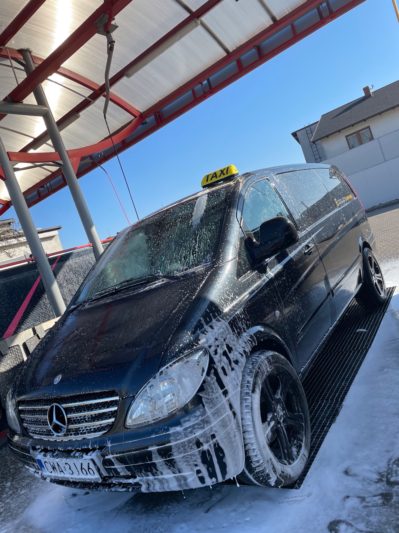 Czarny van Mercedes z żółtym znakiem TAXI na dachu, pokryty pianą z myjni, stojący na metalowej kratce pod zadaszeniem.