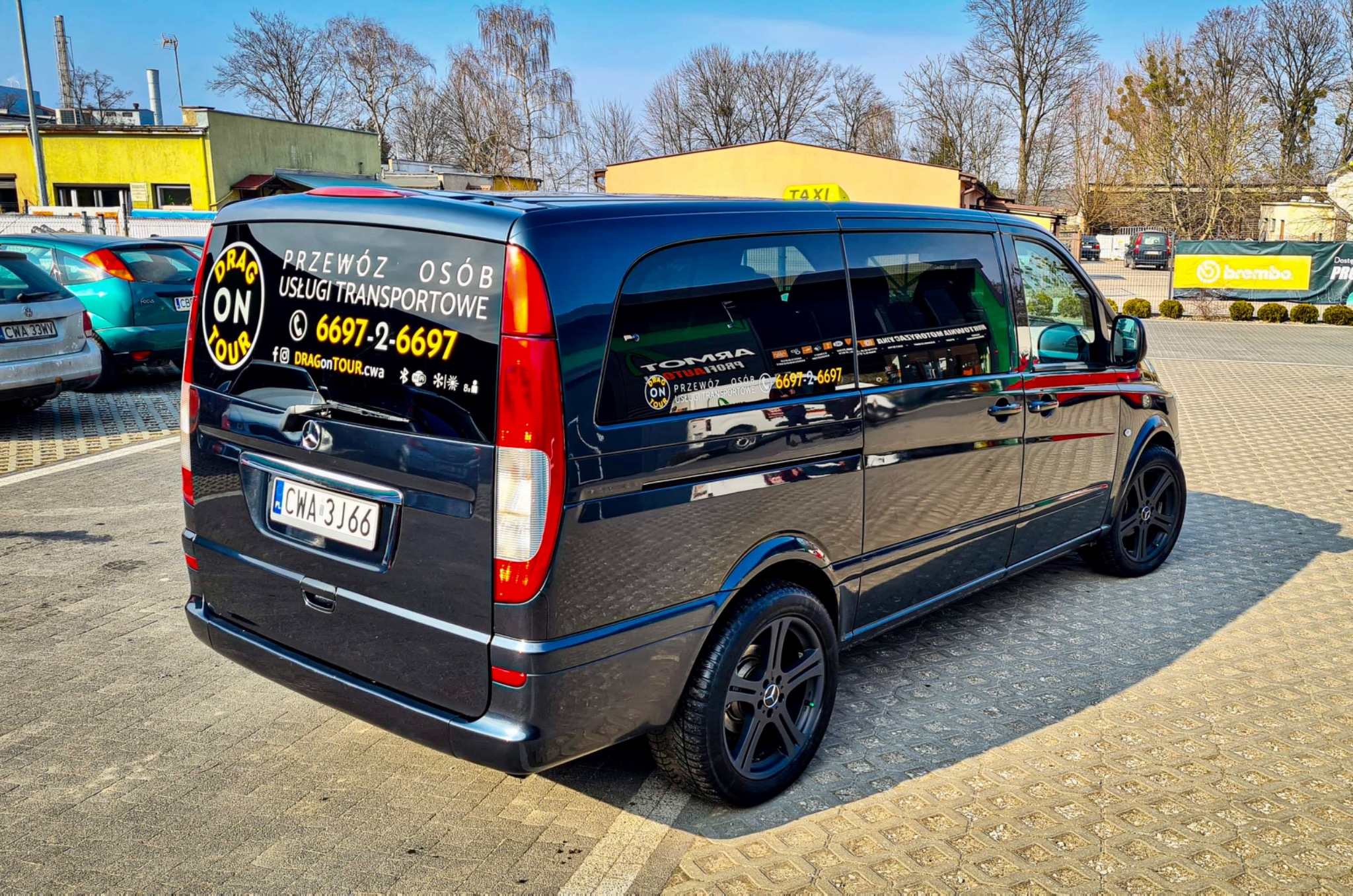 Czarny van Mercedes z naklejkami reklamowymi firmy transportowej na tylnej szybie, zaparkowany na brukowanym parkingu w słoneczny dzień, widoczne numery kontaktowe i adres strony internetowej.