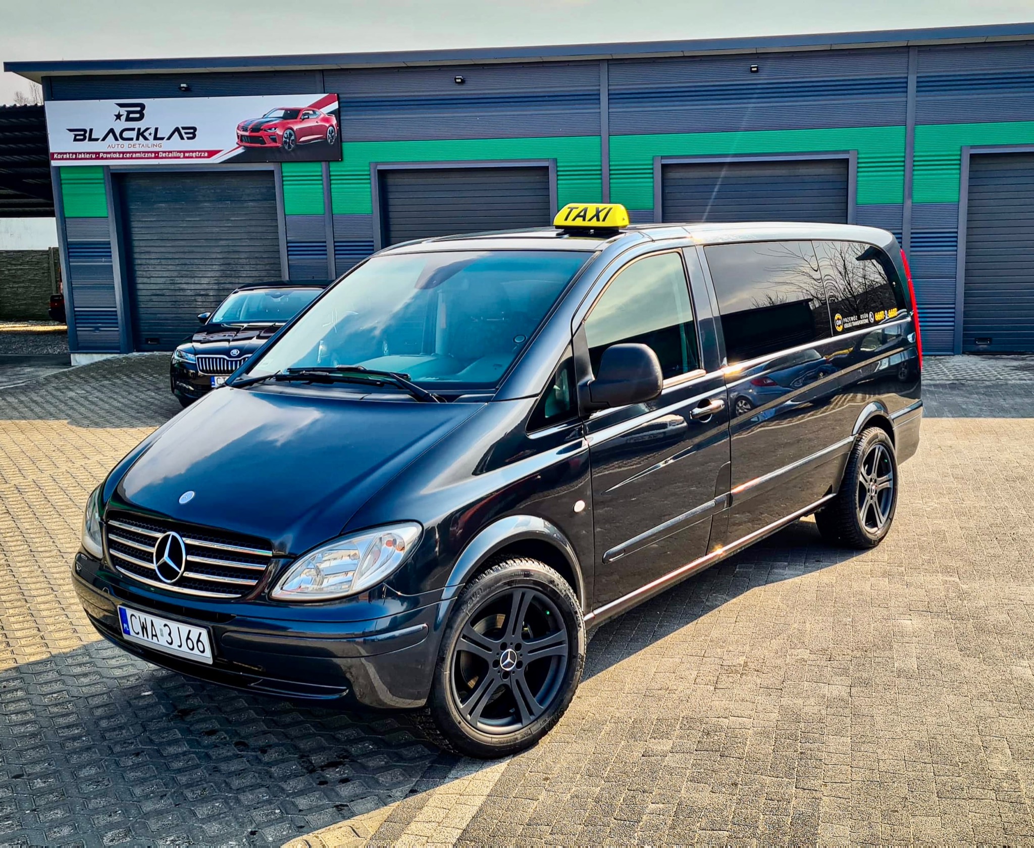 Czarny Mercedes Vito Taxi z żółtym oznaczeniem na dachu, zaparkowany przed budynkiem z logo Black Lab Auto Detailing, obok zaparkowany czarny samochód osobowy.