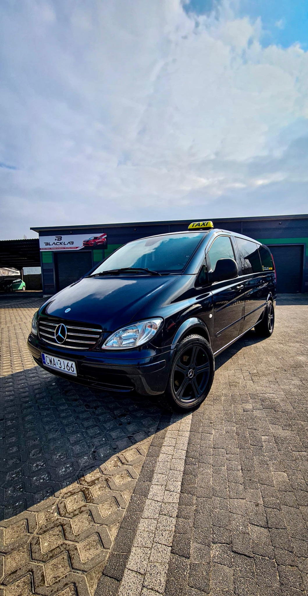 Czarny Mercedes-Benz Vito z żółtym oznaczeniem TAXI na dachu, zaparkowany na brukowanym placu przed budynkiem z logo BLACK LAB, widziany z niskiej perspektywy przy słonecznej pogodzie.