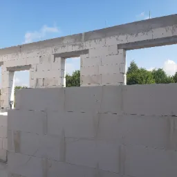 Wznoszenie ścian domu z białych bloczków betonowych, widoczne otwory okienne i betonowy wieniec, na zewnątrz zieleń i błękitne niebo.