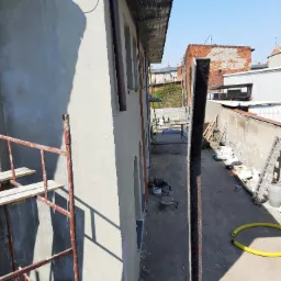 Świeżo otynkowana elewacja budynku z widocznymi rusztowaniami, narzędziami i materiałami budowlanymi na podwórzu, w tle zabudowa miejska.