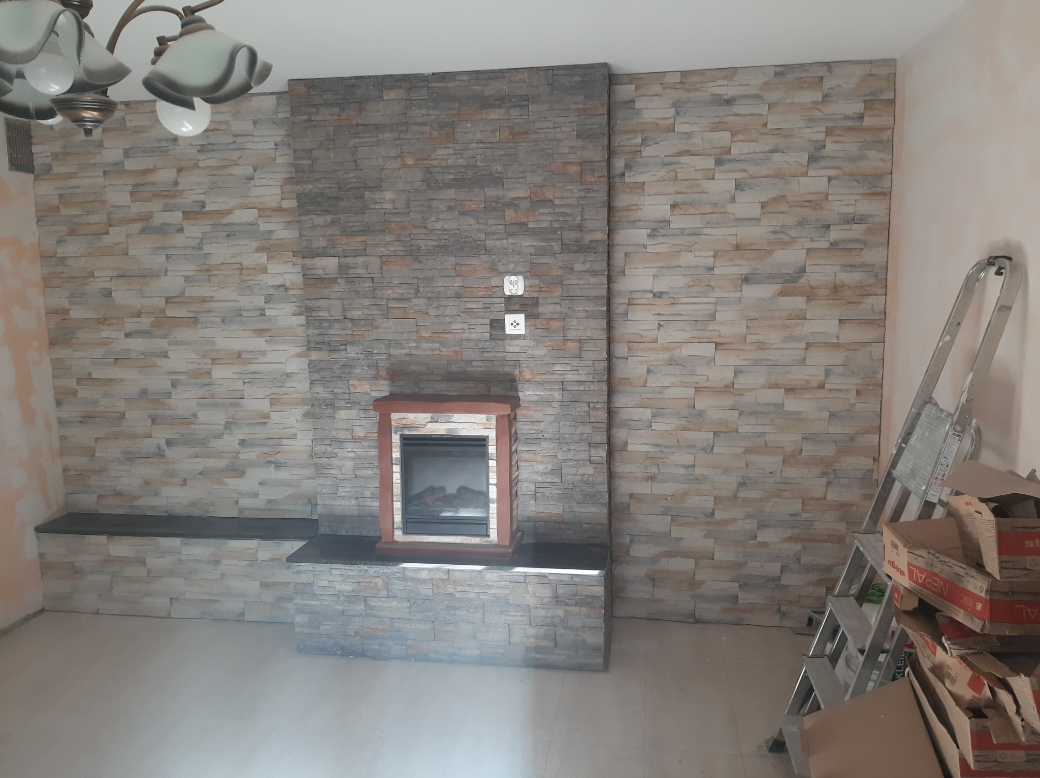 Ściana wykończona kamieniem dekoracyjnym z wbudowanym biokominkiem z drewnianą obudową, widoczna drabina i kartony w rogu pomieszczenia.