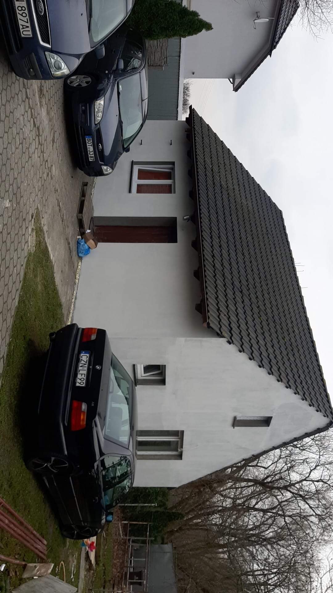 Świeżo otynkowany dom jednorodzinny z ciemną dachówką, widoczne dwa samochody osobowe i fragment podwórka przed budynkiem.