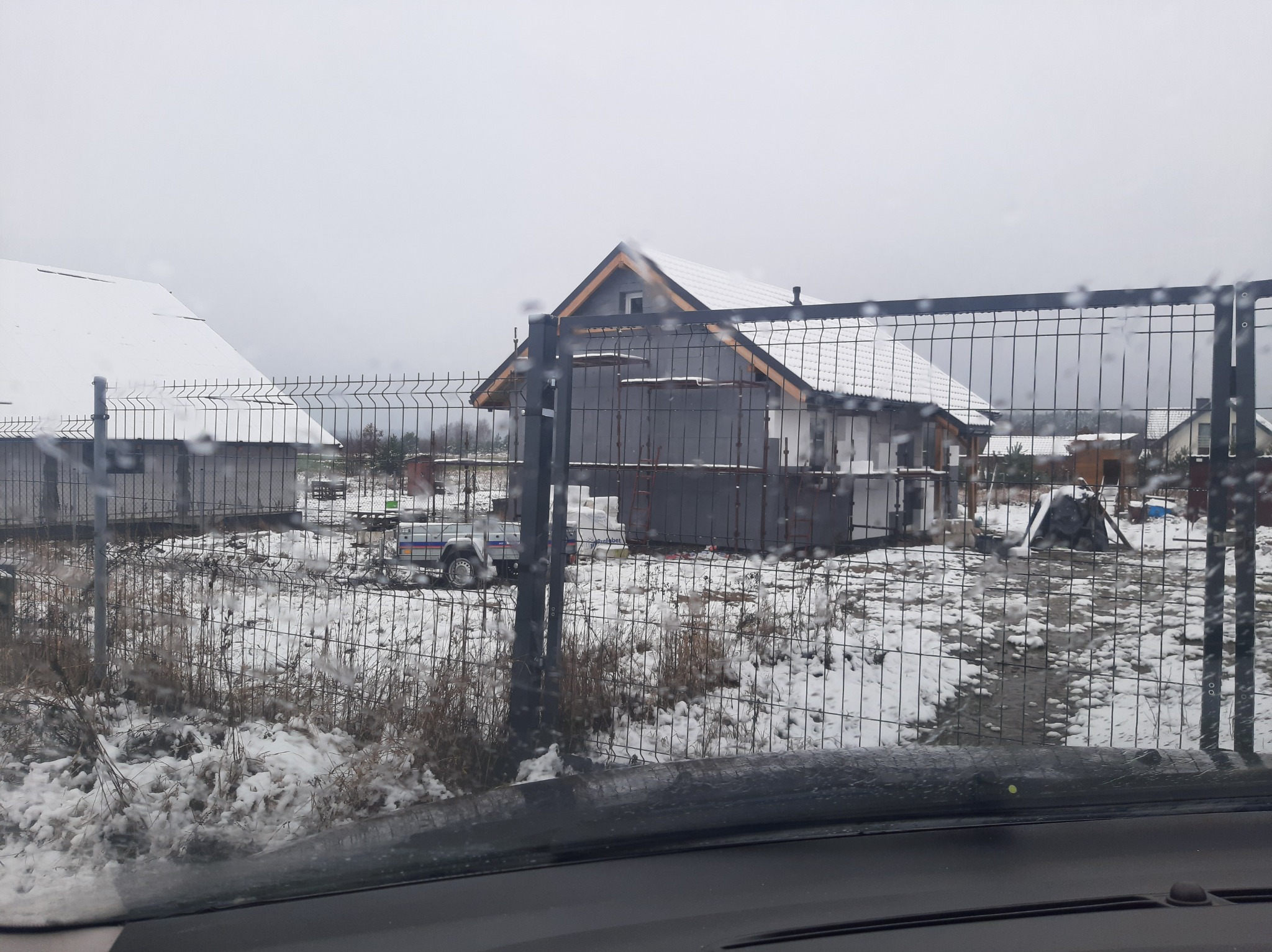 Budowa domu jednorodzinnego w trakcie prac, widok przez mokrą szybę samochodu, ogrodzenie z siatki, teren pokryty śniegiem.