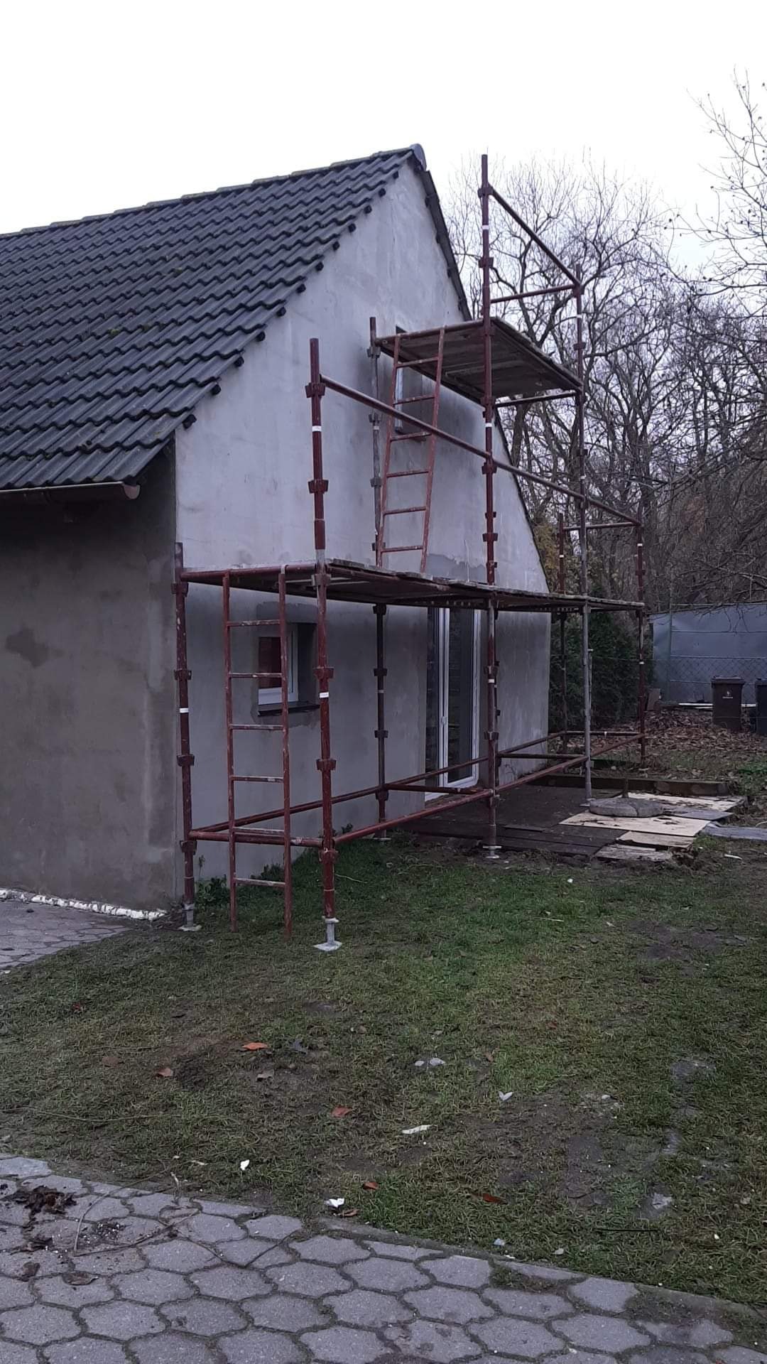 Dom jednorodzinny w trakcie prac elewacyjnych, z widocznymi czerwonymi rusztowaniami i świeżo nałożonym szarym tynkiem. Okna i drzwi zabezpieczone przed zabrudzeniem.