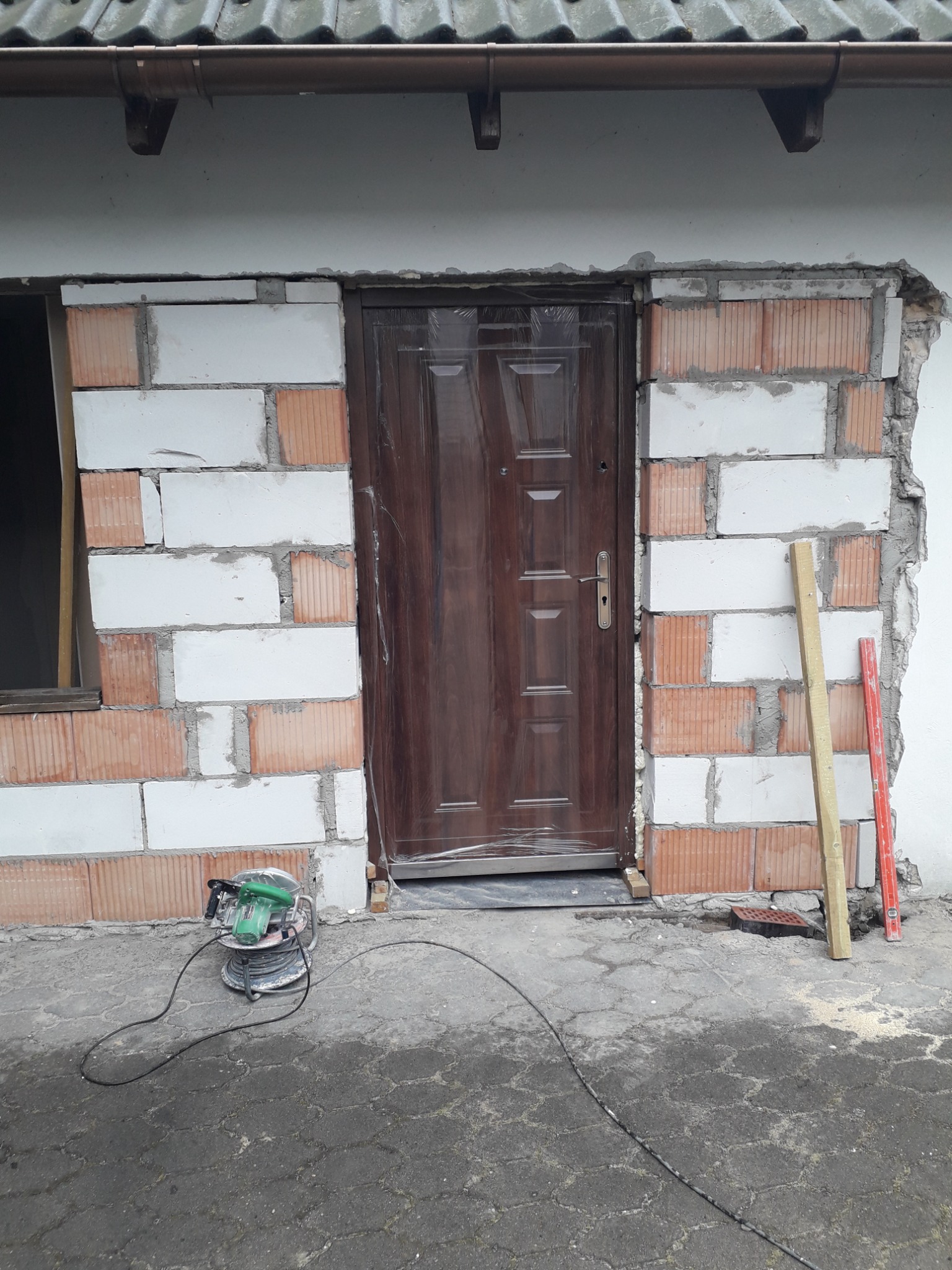 Montaż brązowych drzwi wejściowych w murze z bloczków ceramicznych i białych pustaków, z widoczną przerwą w strukturze ściany i narzędziami budowlanymi obok.