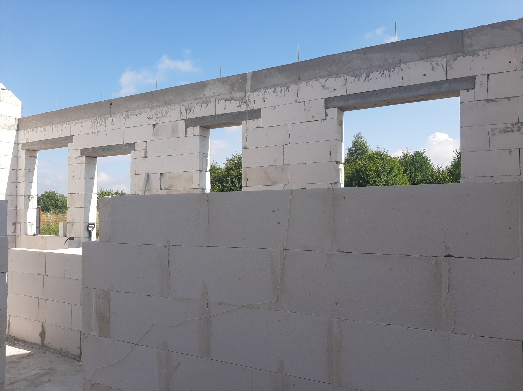 Wznoszenie ścian domu z białych bloczków betonowych, widoczne otwory okienne i betonowy wieniec, na zewnątrz zieleń i błękitne niebo.