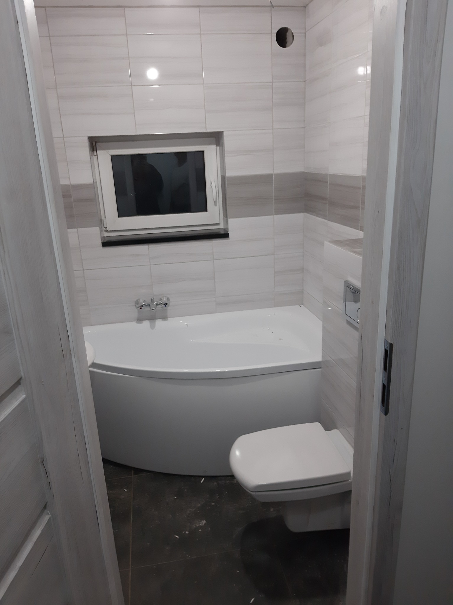 Wykończona łazienka z narożną wanną, podwieszanym WC i małym oknem, widok zza lekko uchylonych, pomalowanych na biało drzwi.