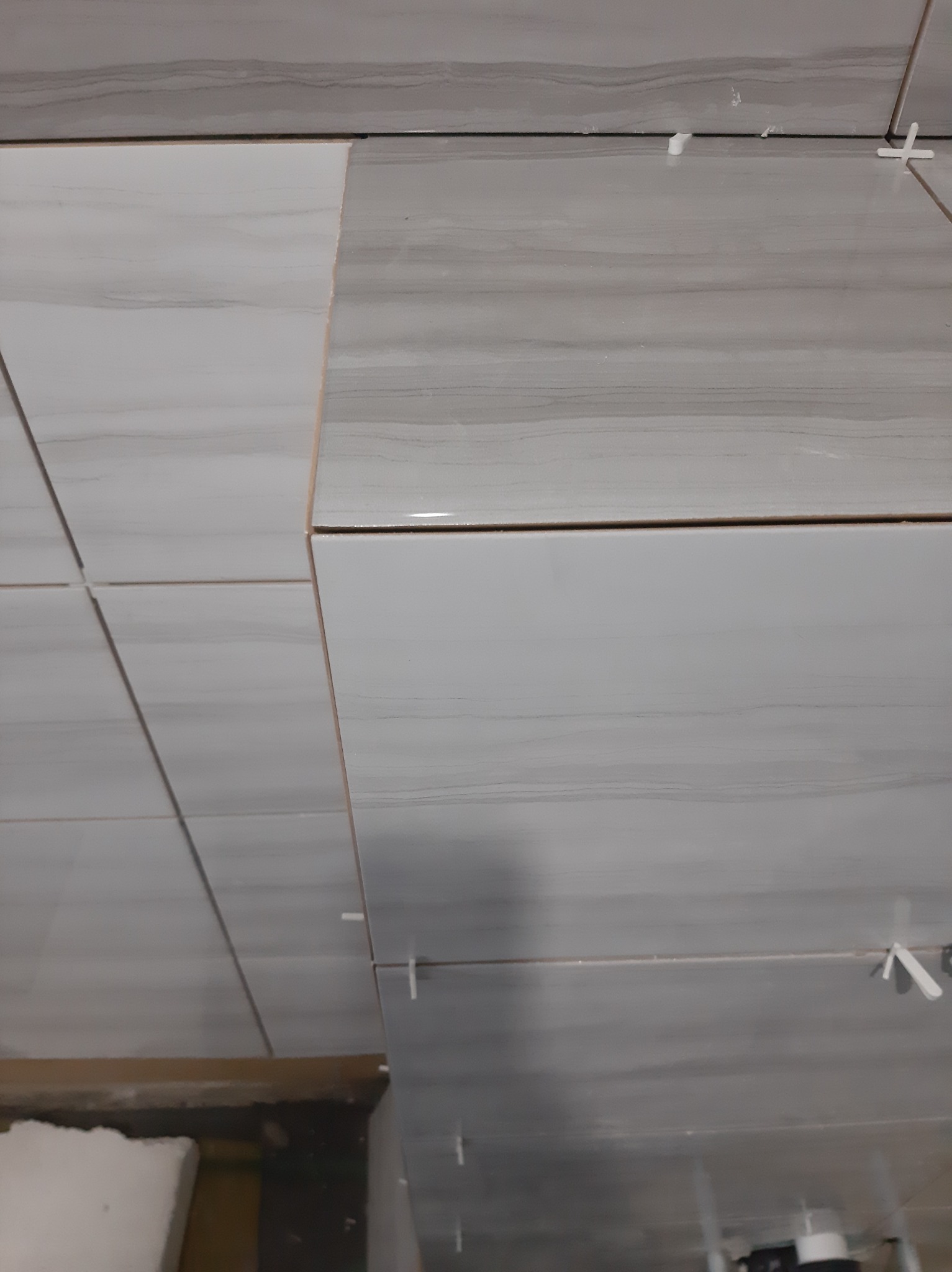 Układanie jasnych, marmurkowych płytek na ścianie z użyciem krzyżyków dystansowych, widoczne niedokończone fragmenty i zaplecze budowy.