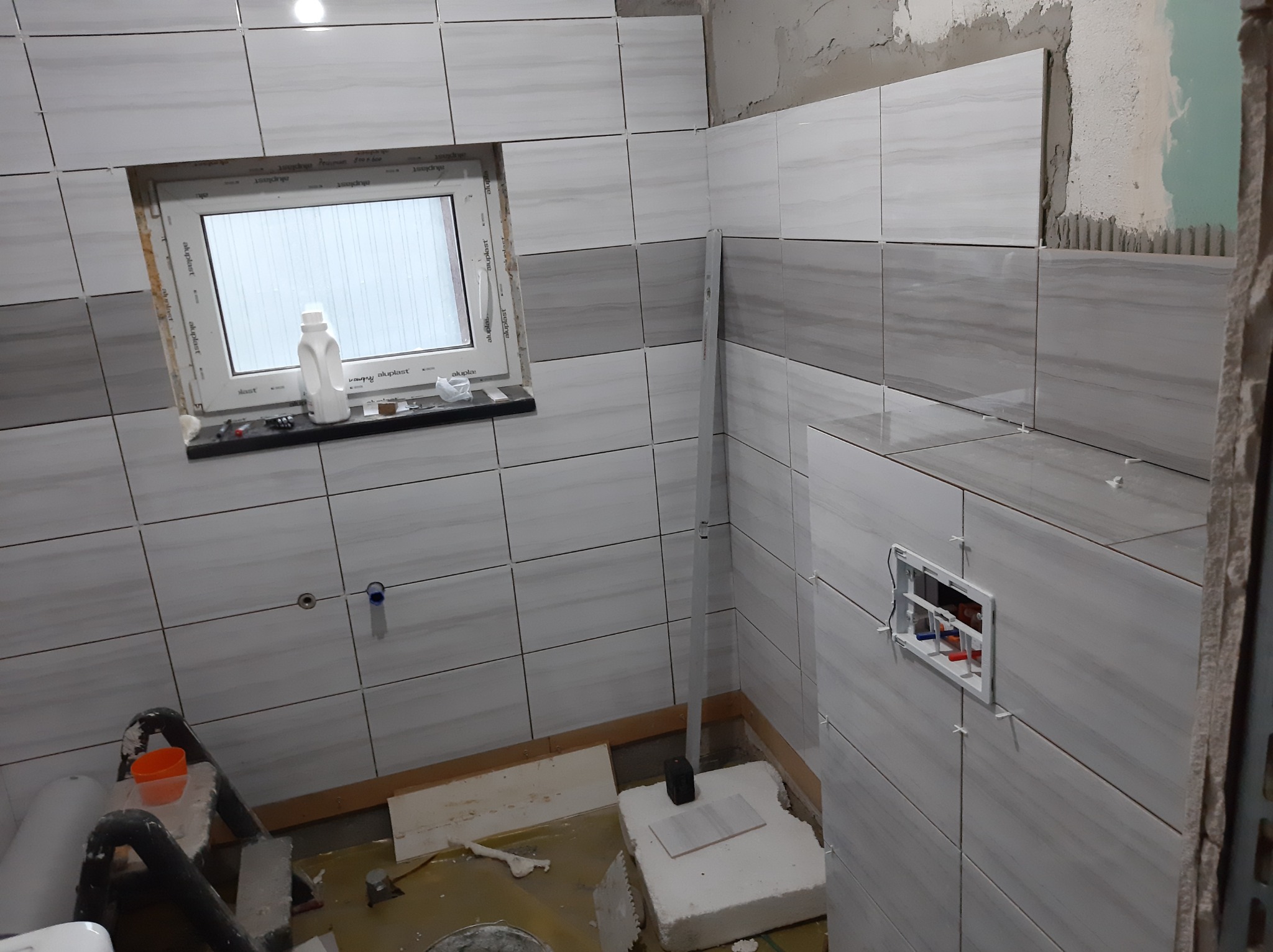 Układanie płytek ceramicznych w łazience, widoczne biało-szare płytki z delikatnym wzorem na ścianach, okno z butelką płynu na parapecie, oraz fragment instalacji wodnej w ścianie, prace w toku.