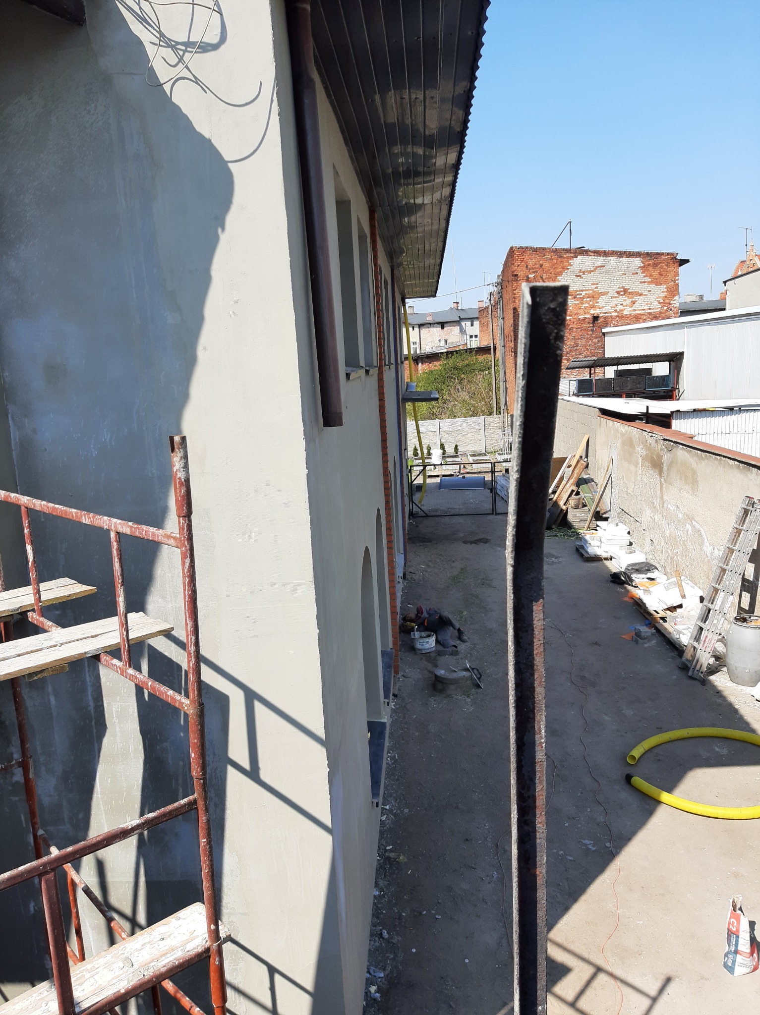 Świeżo otynkowana elewacja budynku z widocznymi rusztowaniami, narzędziami i materiałami budowlanymi na podwórzu, w tle zabudowa miejska.