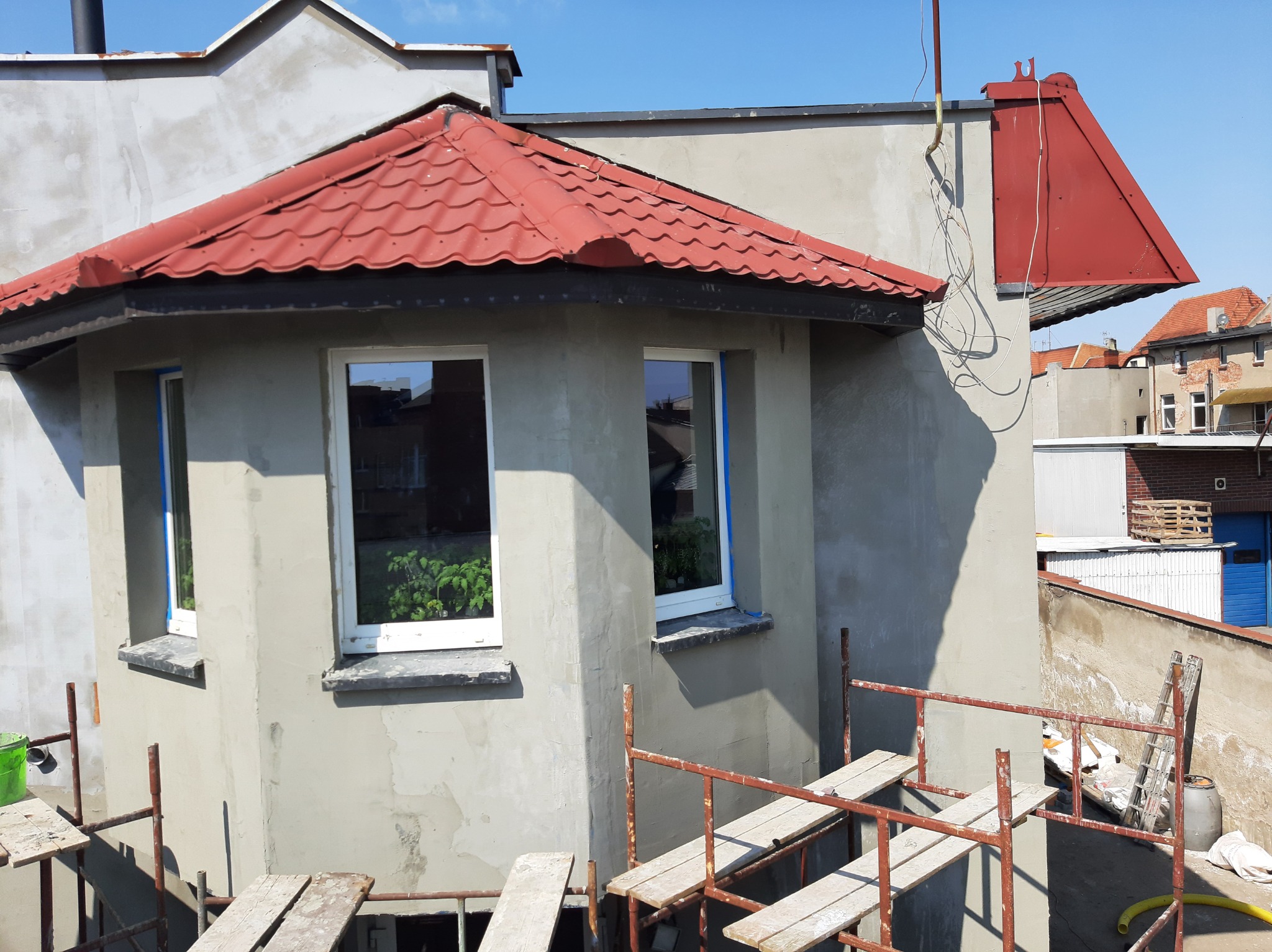 Narożny budynek po ociepleniu ścian zewnętrznych i remoncie elewacji, z nową czerwoną dachówką i oknami z roślinami w tle, widoczne rusztowania.