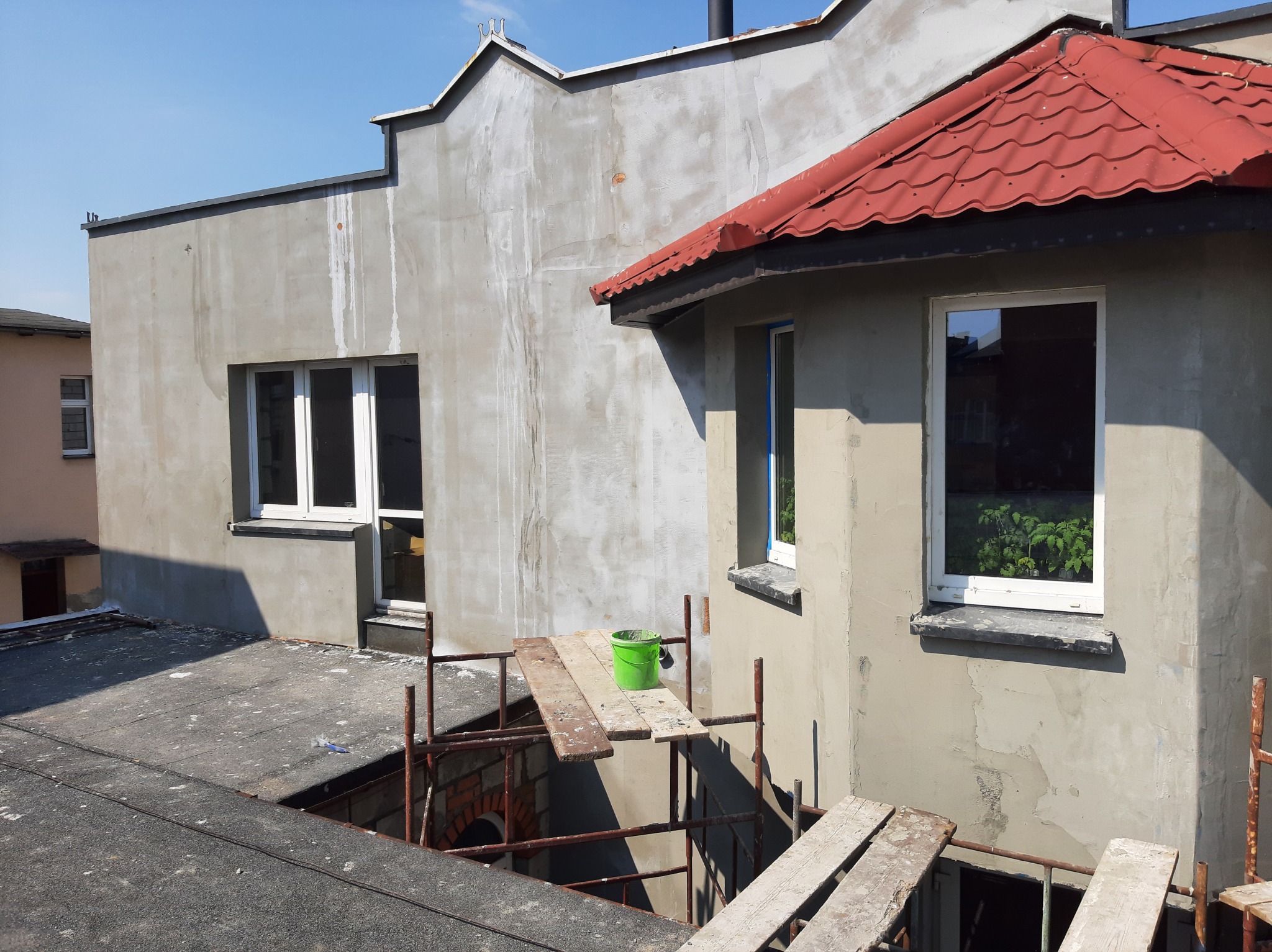 Budynek w trakcie prac elewacyjnych: surowa ściana z oknami, rusztowanie z zielonym wiadrem, czerwony dach nad wykuszem.