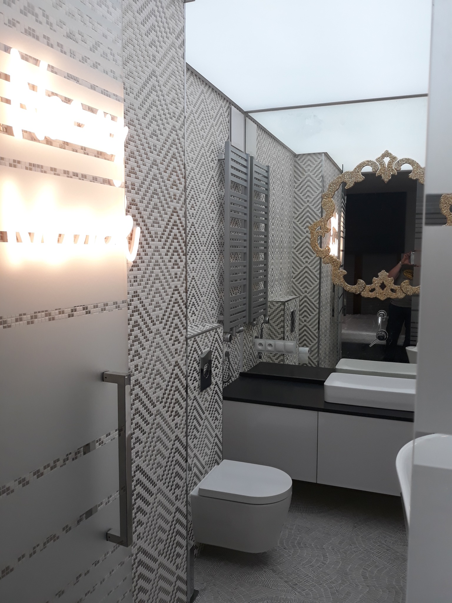 Nowoczesna łazienka z geometryczną mozaiką na ścianach, podświetlanym lustrem, wiszącą toaletą i grzejnikiem drabinkowym, odbicie fotografa w ozdobnym lustrze.
