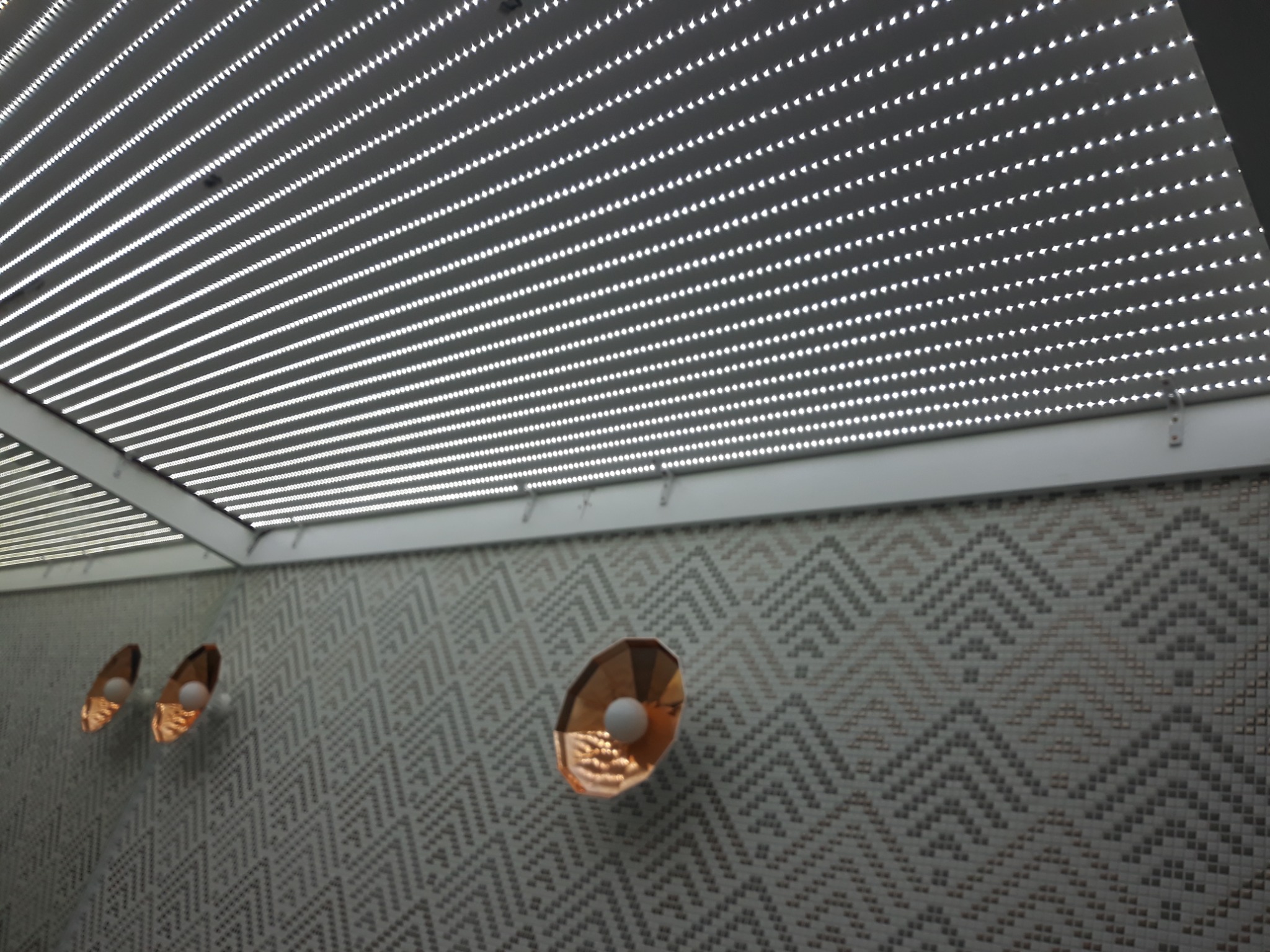 Nowoczesne oświetlenie sufitowe z panelami LED i dekoracyjne lampy ścienne z miedzianymi kloszami na ścianie w geometryczny wzór.