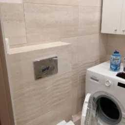 Jasna łazienka z beżowymi płytkami imitującymi kamień, wisząca toaleta z chromowanym przyciskiem spłukiwania, biała pralka Bosch z otwartymi drzwiczkami i szafka wisząca.