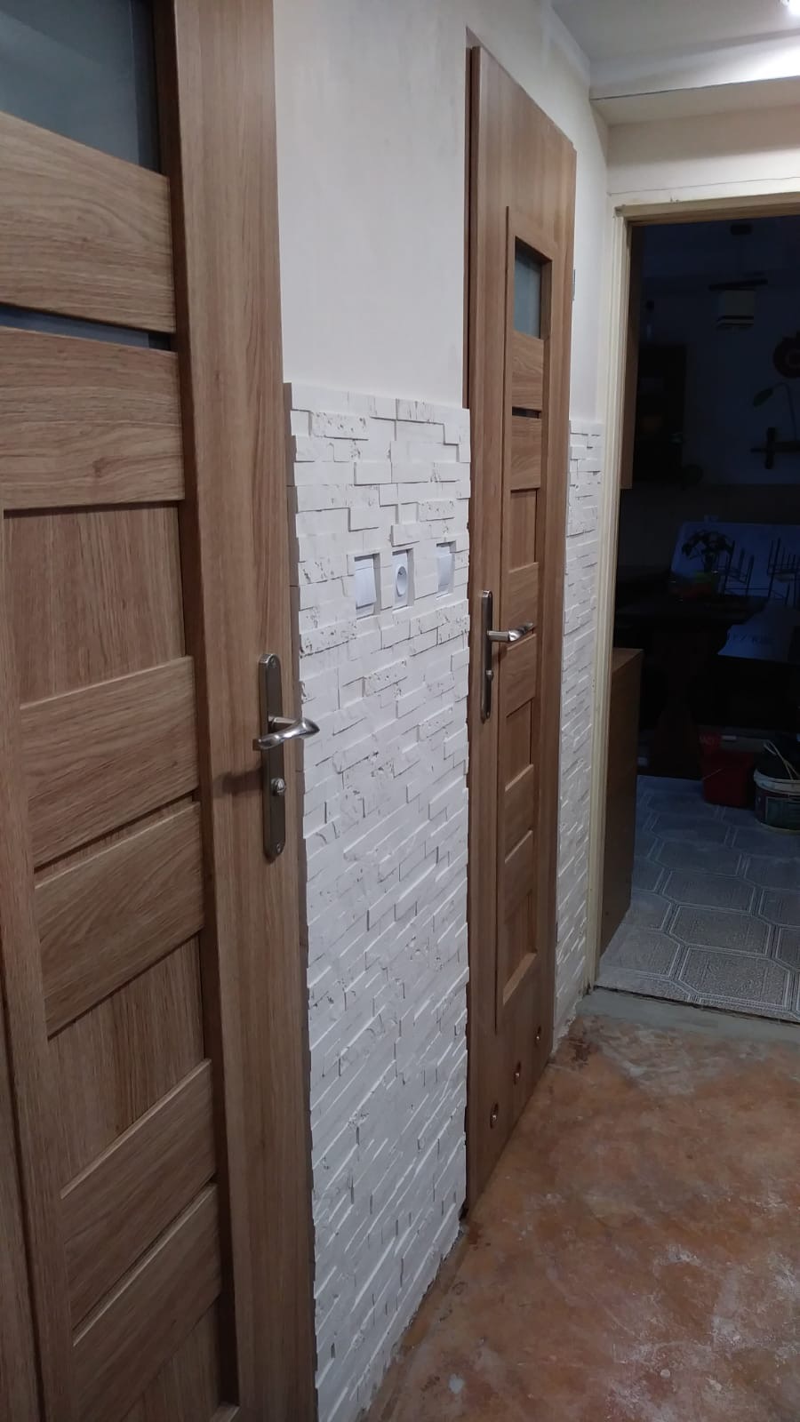 Dwa drewniane drzwi wewnętrzne z metalowymi klamkami, zamontowane obok ściany wyłożonej dekoracyjną białą cegłą, widoczne w korytarzu z fragmentem podłogi.