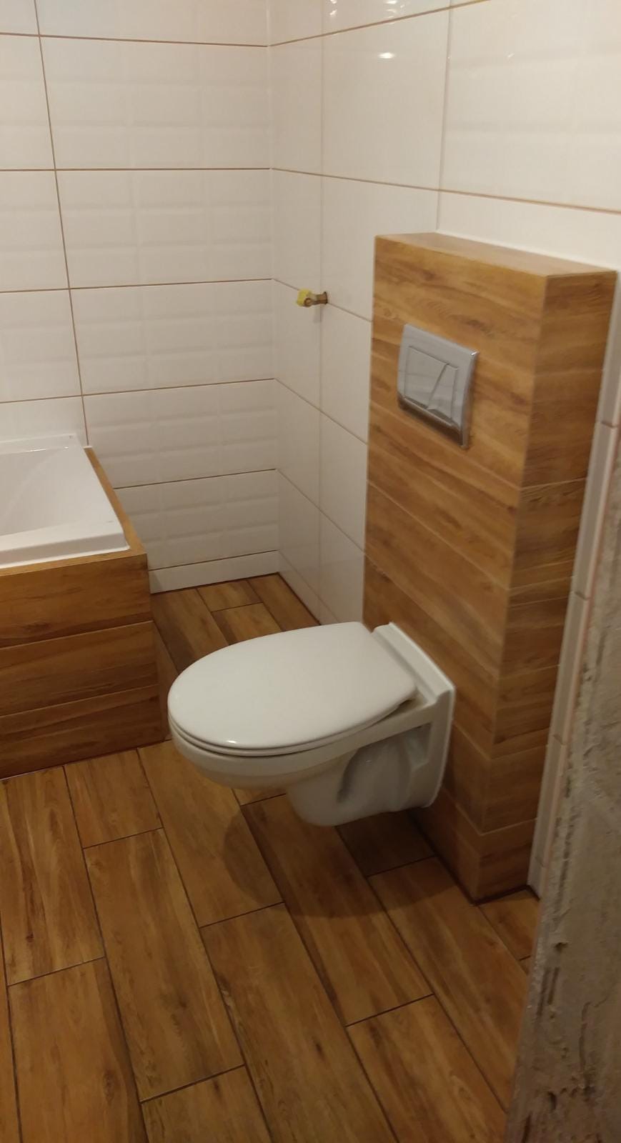 Nowoczesna łazienka z podwieszaną toaletą obudowaną drewnopodobną płytą, białe płytki na ścianach i drewnopodobna podłoga, widoczny fragment obudowy wanny.