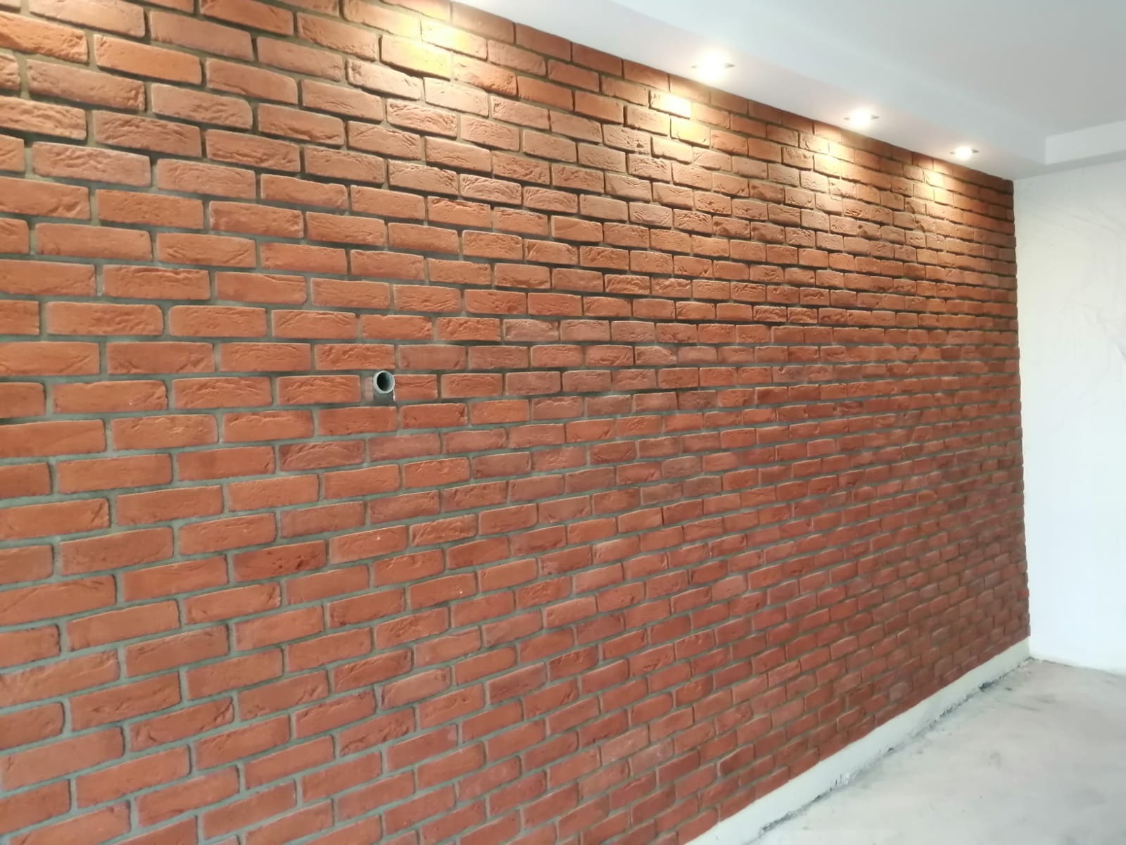 Ściana wykończona czerwoną cegłą dekoracyjną z widocznym otworem na instalację elektryczną i oświetleniem punktowym na suficie.