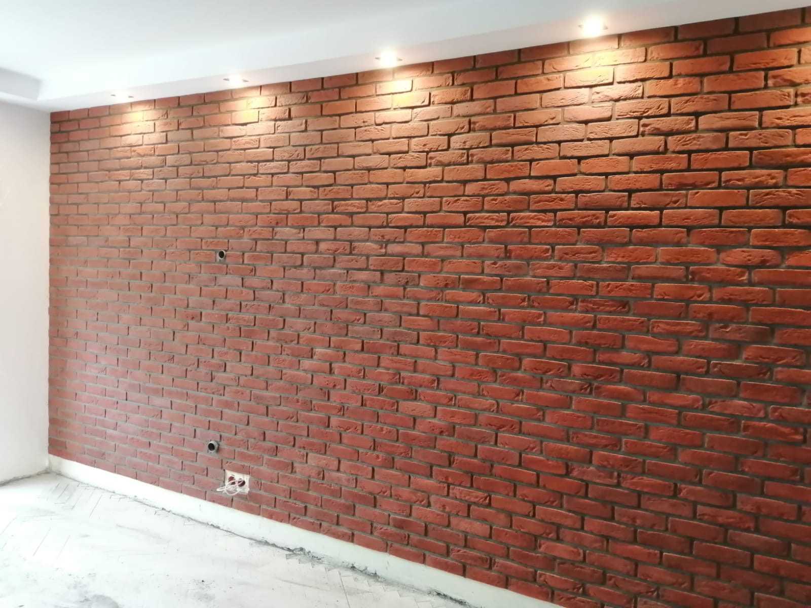 Ściana z czerwonej cegły dekoracyjnej w jasnym wnętrzu, oświetlona punktowymi lampami sufitowymi, z widocznymi gniazdkami elektrycznymi i otworami na kable.