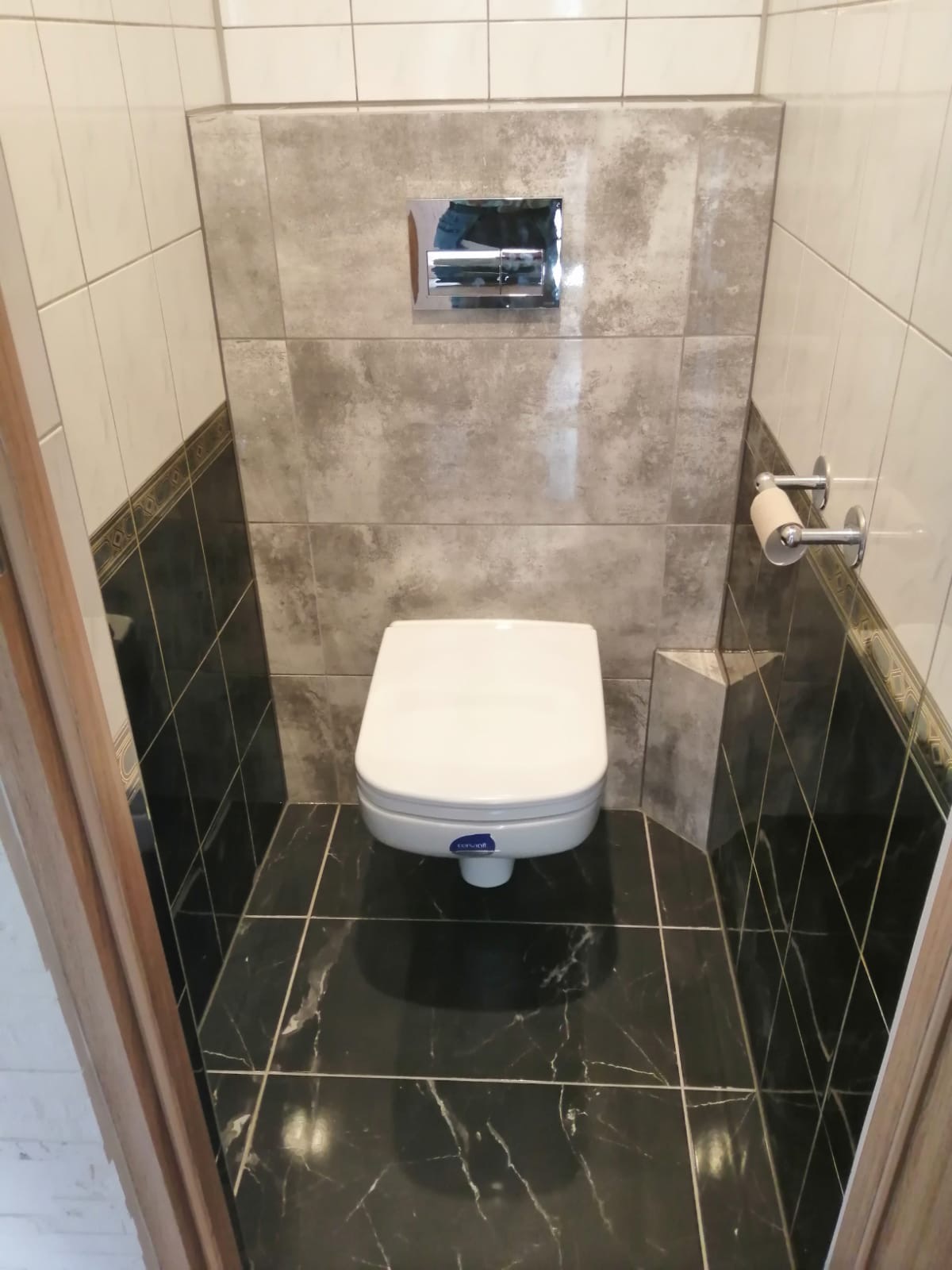 Kompaktowa toaleta podwieszana z chromowanym przyciskiem spłukującym, obudowana płytkami imitującymi kamień w kolorze szarości i czerni z dekoracyjnym paskiem, oraz uchwyt na papier toaletowy.