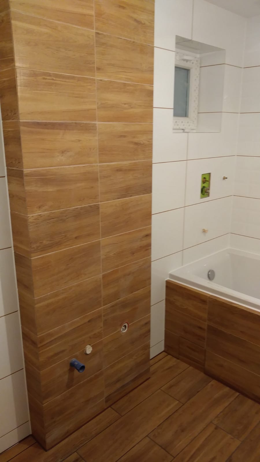 Nowoczesna łazienka z drewnopodobnymi płytkami na ścianach i podłodze, biała wanna z obudową z płytek w kolorze drewna, małe okno z białym obramowaniem, widoczny fragment instalacji hydraulicznej.