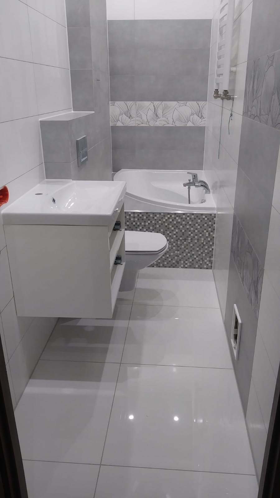 Nowoczesna łazienka w odcieniach szarości i bieli z wiszącą szafką umywalkową, toaletą i wanną z mozaiką, wykończona błyszczącymi płytkami podłogowymi.