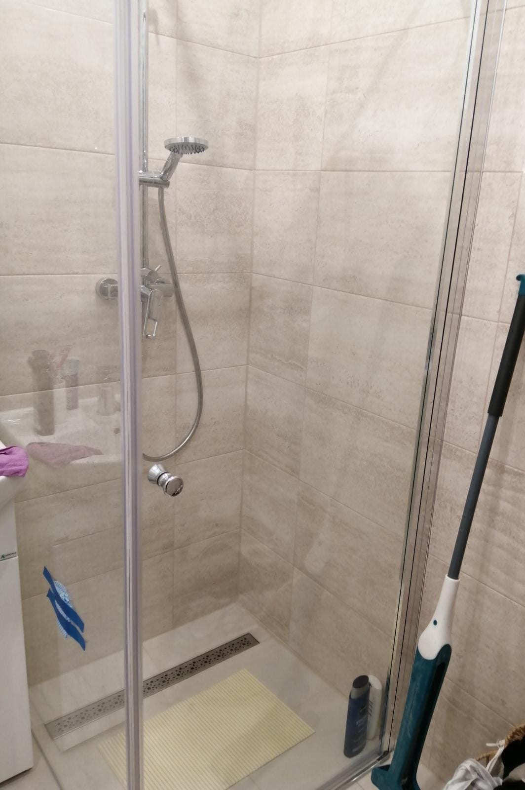 Wykończona kabina prysznicowa z odpływem liniowym, jasnymi płytkami imitującymi kamień i mopem parowym w rogu.