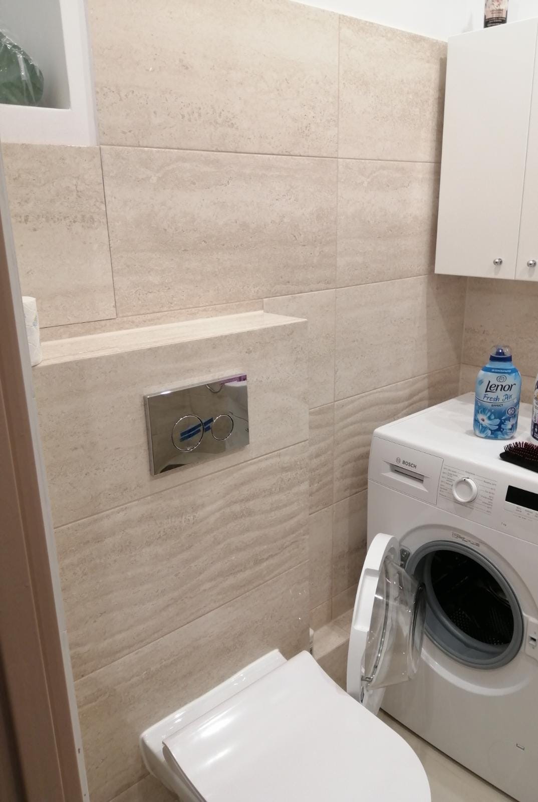 Jasna łazienka z beżowymi płytkami imitującymi kamień, wisząca toaleta z chromowanym przyciskiem spłukiwania, biała pralka Bosch z otwartymi drzwiczkami i szafka wisząca.