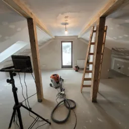 Poddasze w trakcie szpachlowania i przygotowania do malowania, widoczne drewniane belki konstrukcyjne, drabina, szpachlowane ściany, lampa budowlana na statywie oraz szlifierka do gładzi...