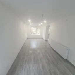 Jasne, przestronne wnętrze z panelami podłogowymi w odcieniach szarości. Białe ściany, okno z widokiem na zieleń i grzejnik. Sufit z punktowym oświetleniem LED.