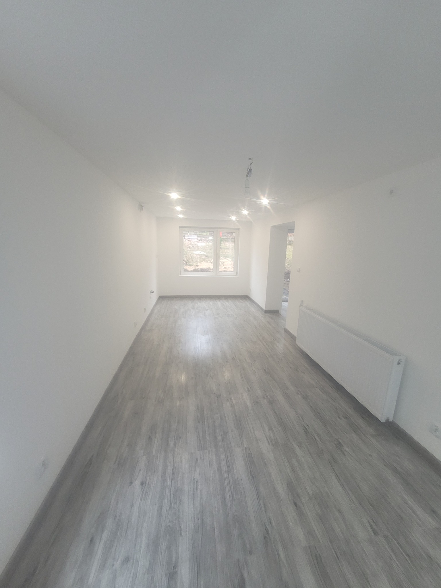 Jasne, przestronne wnętrze z panelami podłogowymi w odcieniach szarości. Białe ściany, okno z widokiem na zieleń i grzejnik. Sufit z punktowym oświetleniem LED.